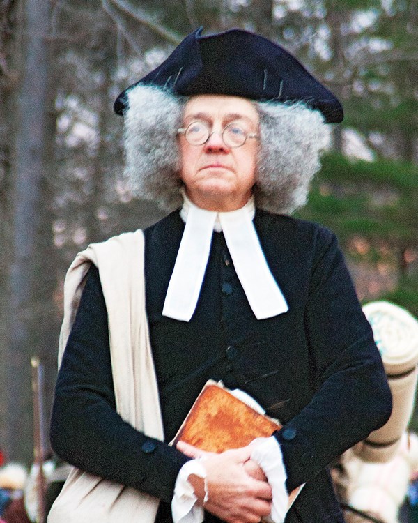 Man in 18th century minster's clothing, black coat, gray wig, white ministerial collar.