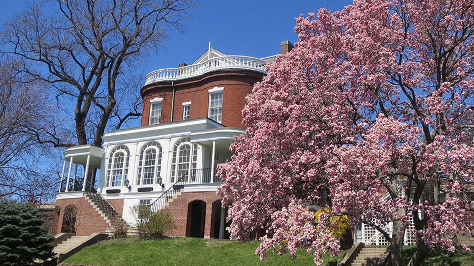 Commandant House, a red brick and white wood Federalist Style structure next to a blooming tree.