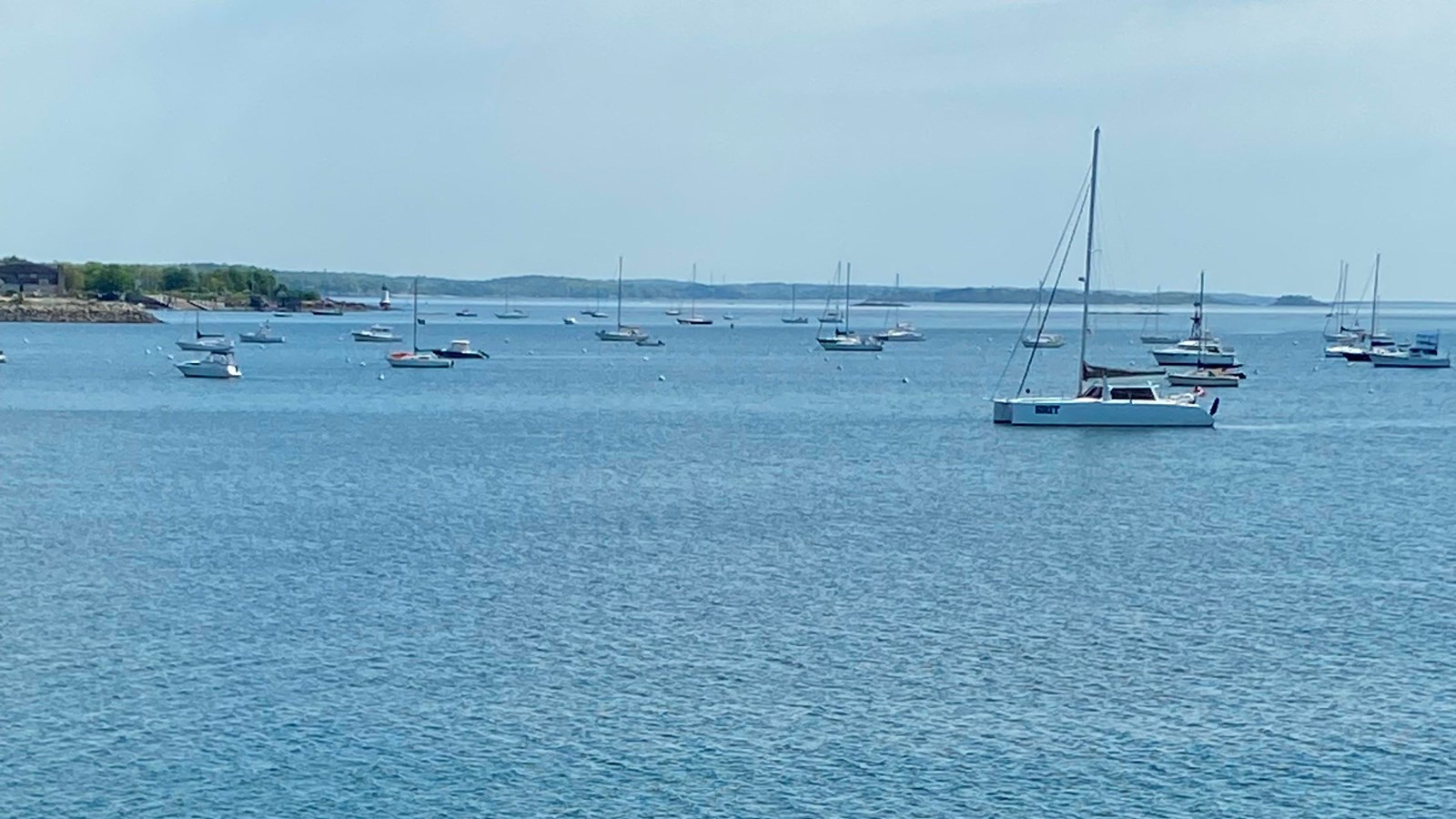 Bright blue waters with many sailing vessels anchored and land jutting out in the harbor on the left