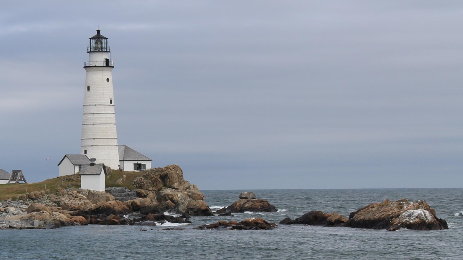 A tall white lighthouse with smaller white buildings at its foot along a rocky coastline.