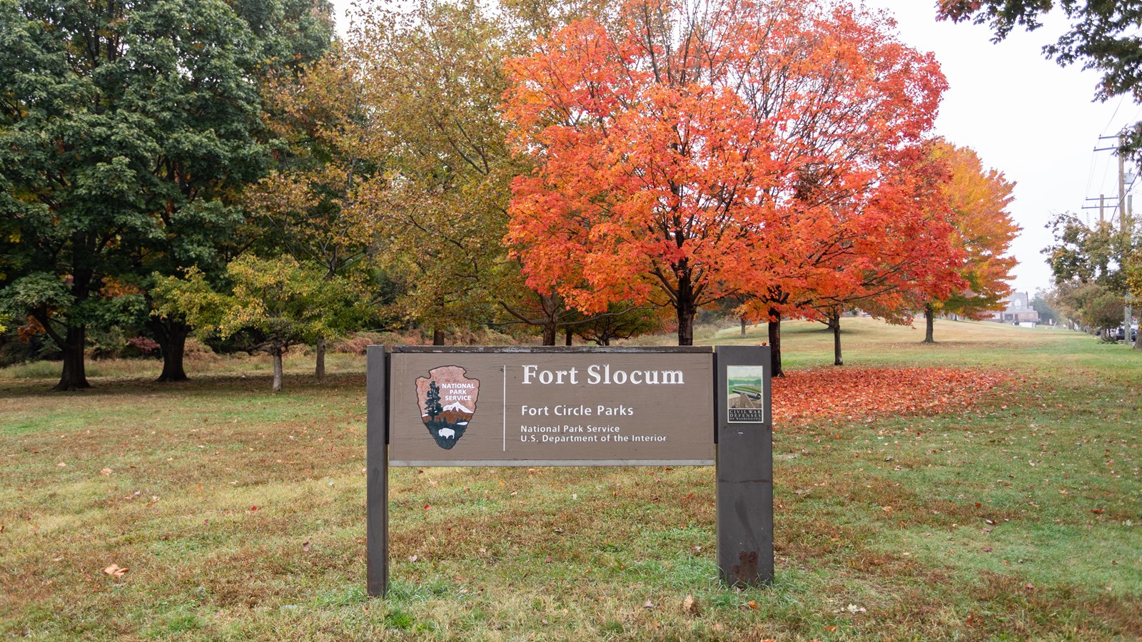 A sign with the words Fort Slocum in front of a tree with red leaves. 
