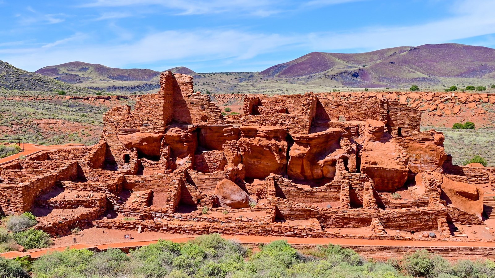 A large red sandstone pueblo with many visible rooms in a desert grassland with mountains behind.  