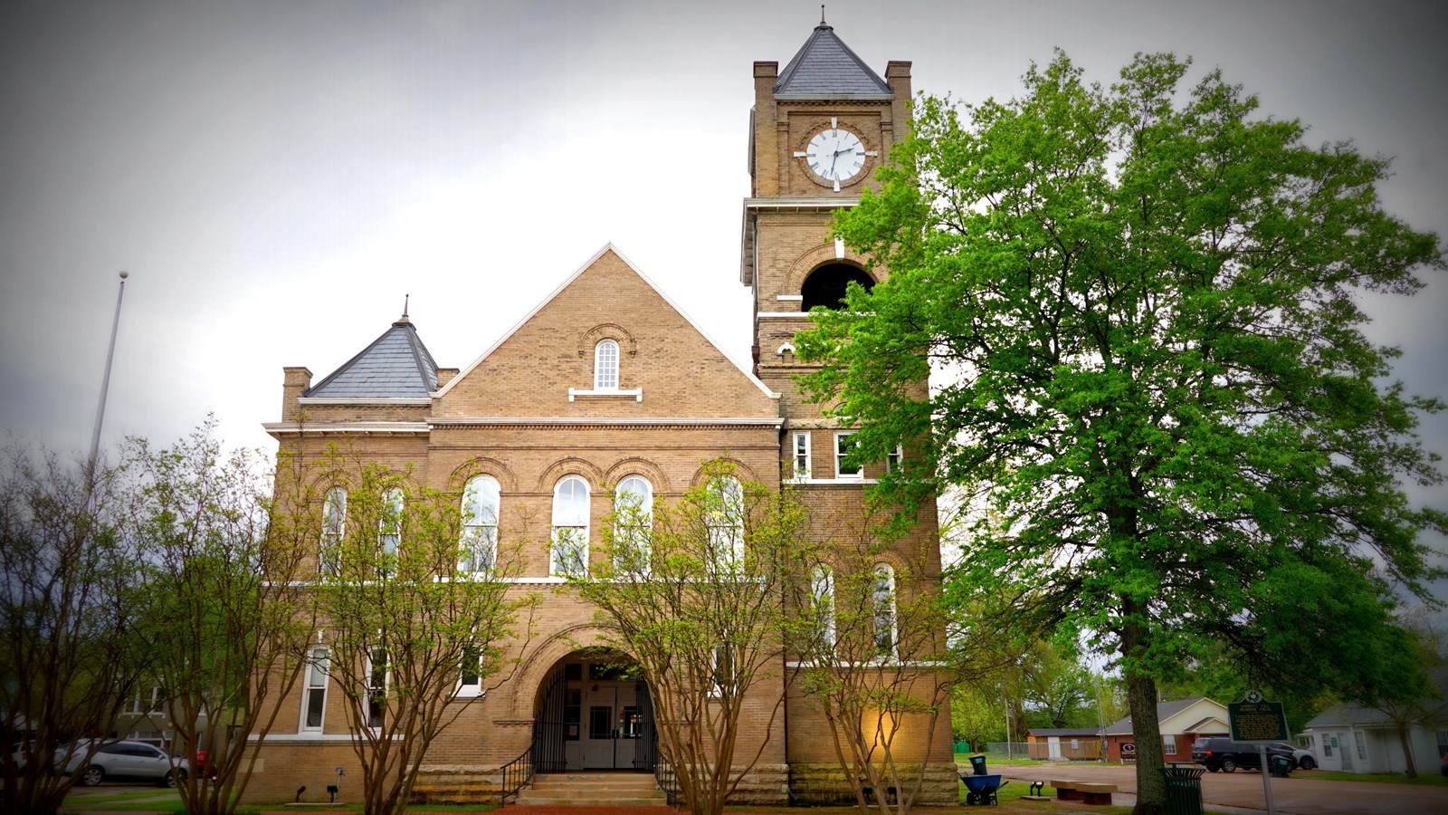 Front of a two-story brick courthouse with a clock tower. A tree stands in front. 