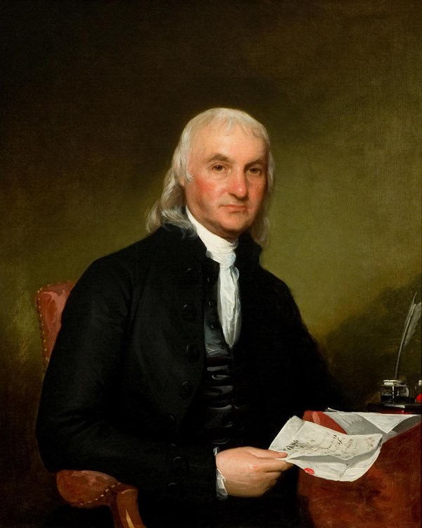A seated man with long white hair wearing a dark suit and holding papers.
