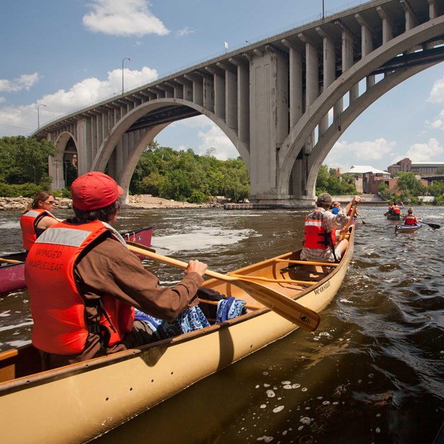 Canoeists paddle a large river beneath a bridge.