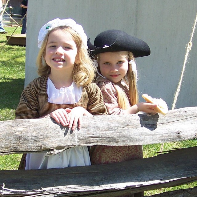 Two small girls with light blonde hair dressed in colonial attire lean on a wooden fence.