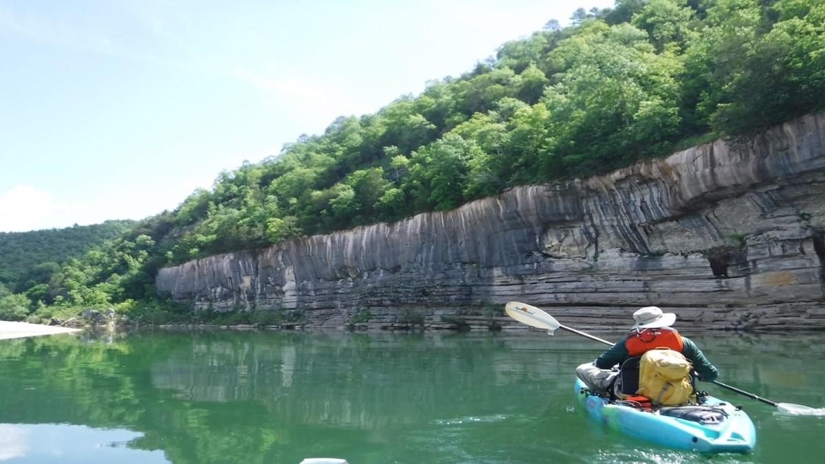 A paddler in a teal kayak floats on blue-green water as they pass by a gray bluff and green hillside