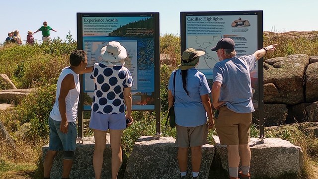 Two groups of people standing in front of vertical orientation waysides on mountain summit