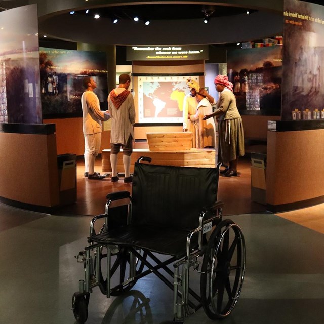 A wheelchair in front of a burial scene with 5 life-sized touchable figures and clothing.