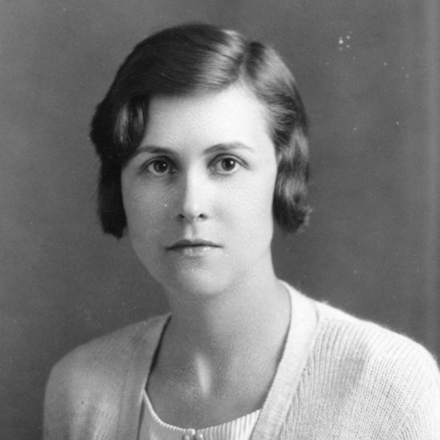 Black and white image of a woman in a white blouse and white sweater looking intently at camera
