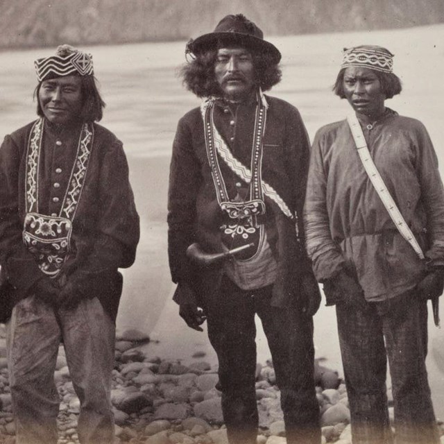 Three men standing by the side of a river. Black and white photo.