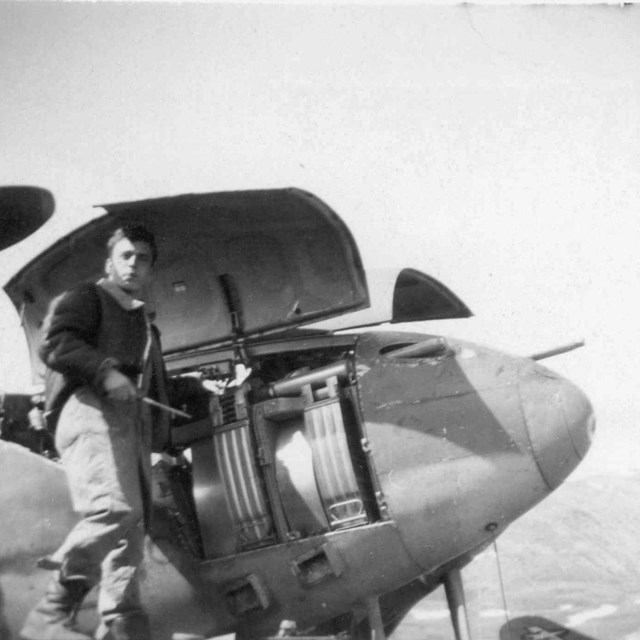 Black and white photo of man holding tools in front of nose of airplane. 
