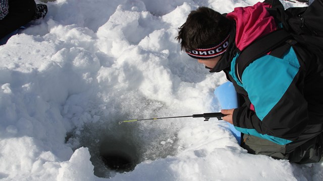 Close-up of kid with fishing pole laying down on ice looking down a hole in the ice.