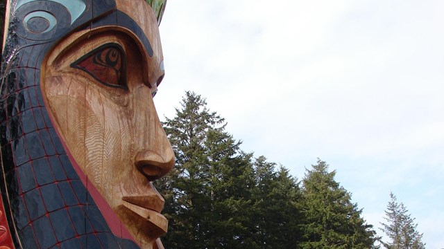 Close-up of carved totem pole with cloudy sky, green trees, and building in background.