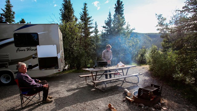 Campsite; back end of RV, person in camp chair, person at picnic table, campfire.