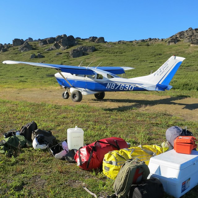 A small plane is parked at the end of a gravel air strip. A pile of gear is near the airstrip.