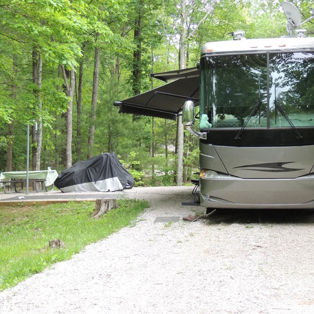 Large RV sits upon a gravel lot near a tent pad with picnic table.