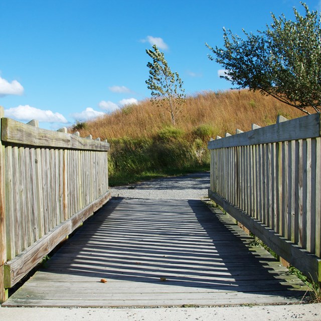 A wooden bridge leads to a gravel trail. Tall grass on a hill in the background.