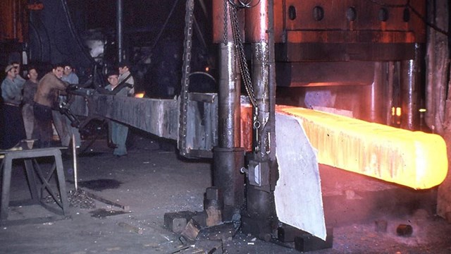 An anchor being forged in a shop with the end of metal bright yellow & men working at the other end.