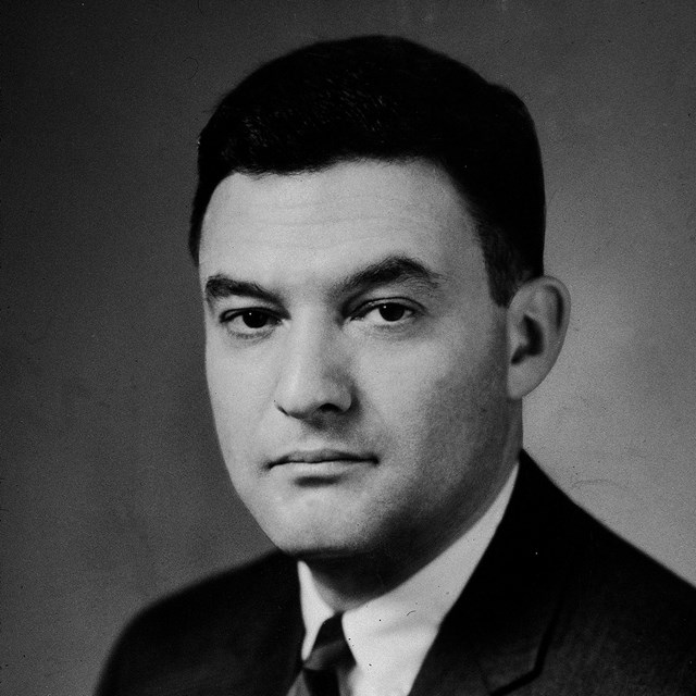 Jack Greenberg poses for a head and shoulders portrait in a black suit and striped tie.