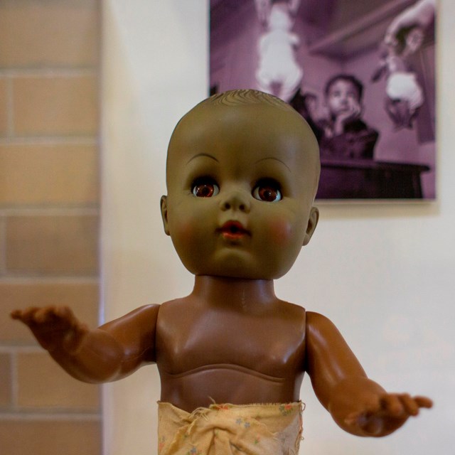 Brown doll in front of a photo of an African American child choosing between a while and brown doll.