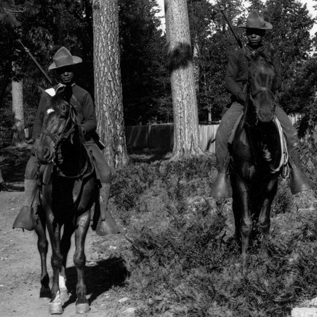 5 soldiers on horseback wearing hats with rifles in front of trees.