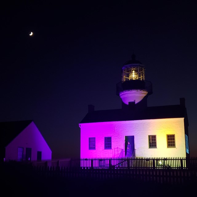 White lighthouse half lit up in purple light and other half lit with yellow lights. Yellow light in 