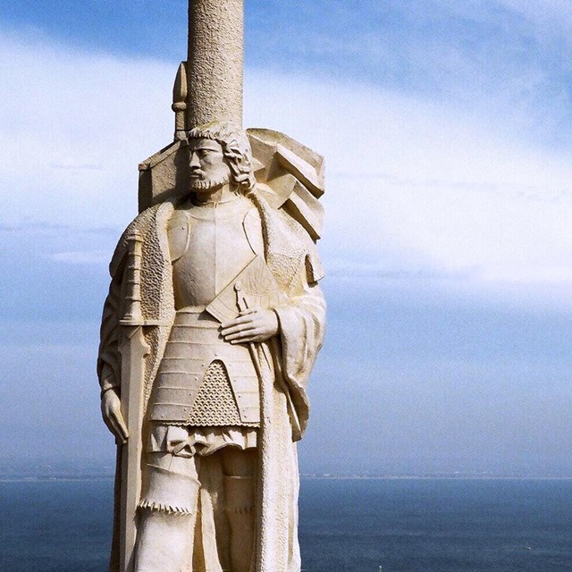 White stone statue of a man with cape. Blue ocean and blue sky with white clouds behind it.