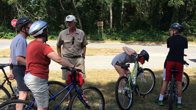 A man in a khaki park service volunteer shirt talks to bicyclists wearing helmets on a paved trail.