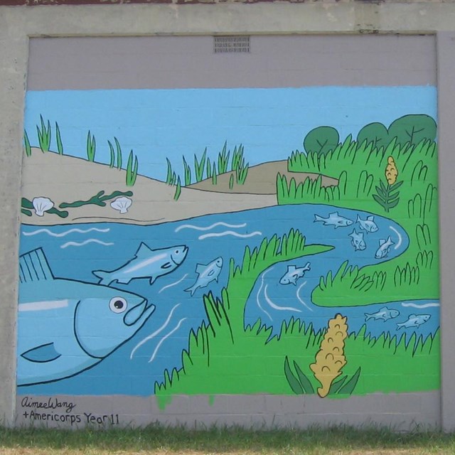 Two side-by-side painted murals on the side of a cinder block building depict o river scene.