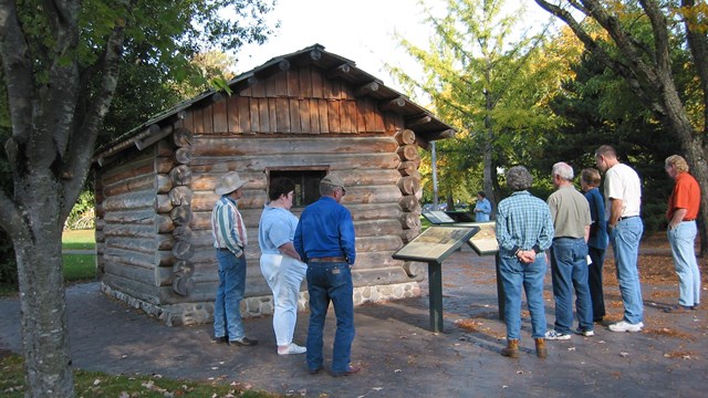 A group of people stands around a wayside exhibit next to a log cabin.