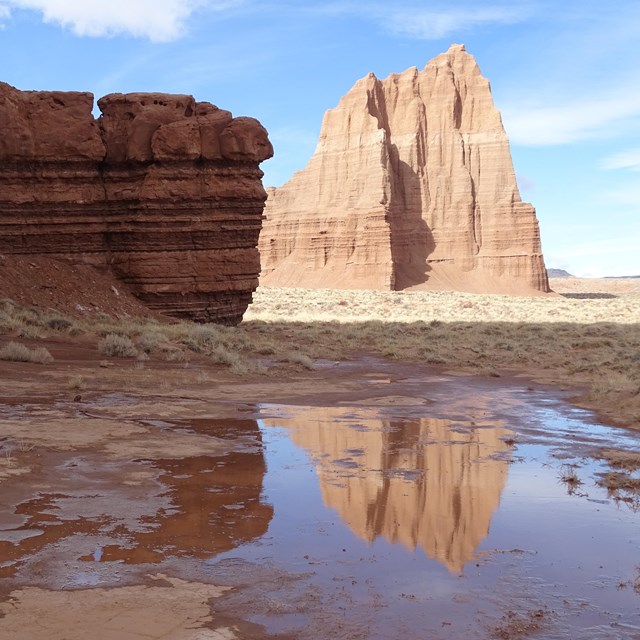 Large red, triangular monolith, reflected in a puddle, with blue sky above, and red rock to the side