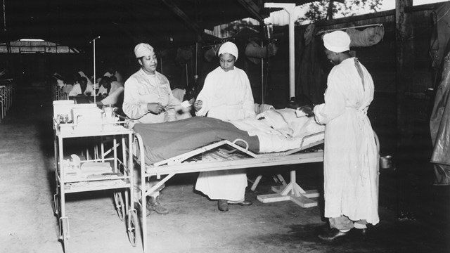 Three Black nurses in white surgical gowns tend to a man on a hospital bed. 