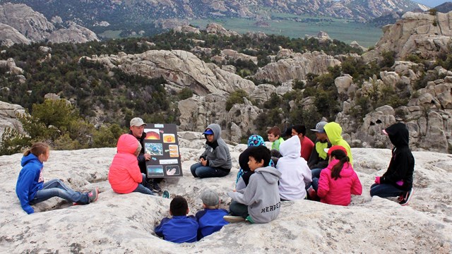 A group of kids sitting on rock slab listening to a Ranger with a poster board at City of Rocks.