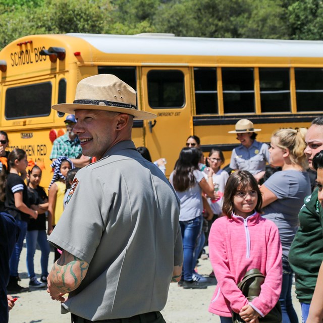 A ranger smiles while surrounded by children in front of a school bus