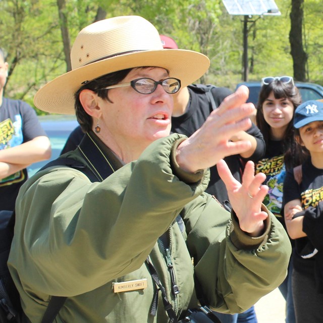 A park ranger stands in front of a group of kids while pointing in the distance, trees in background