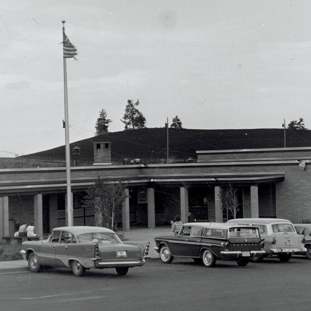 black and white photo of a brick building with 1950s era cars parked in front