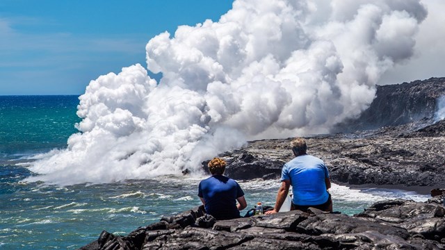 Two hikers sitting watching a volcano next to the ocean erupt