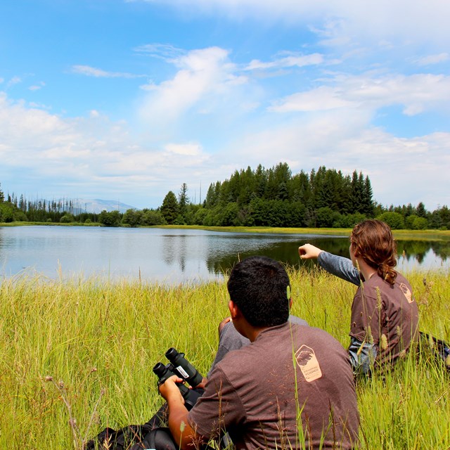 Two high school students sit in tall grass near edge of lake; one points toward lake.