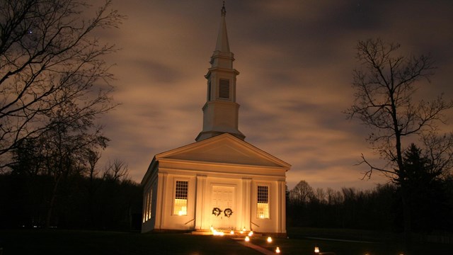  It is a cloudy night. Tree silhouettes surround a white church, lit by lanterns from below.