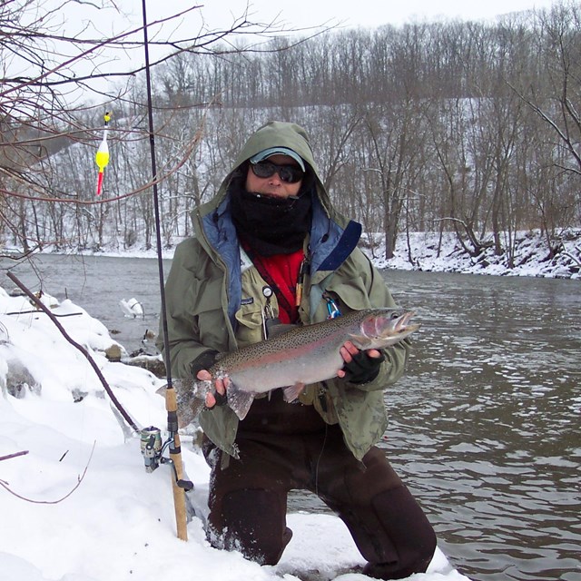 Person in a winter coat holds a fish with both hands next to a river, the banks covered in snow.