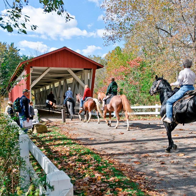 Five people astride horse make their way toward and through a red covered bridge.