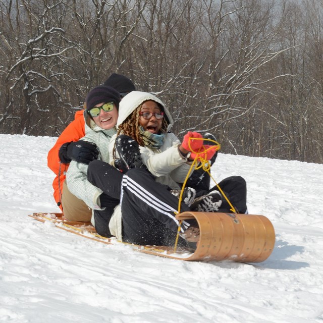 Three people in winter coats and gloves smile as they ride down a snowy hill on a wooden toboggan.