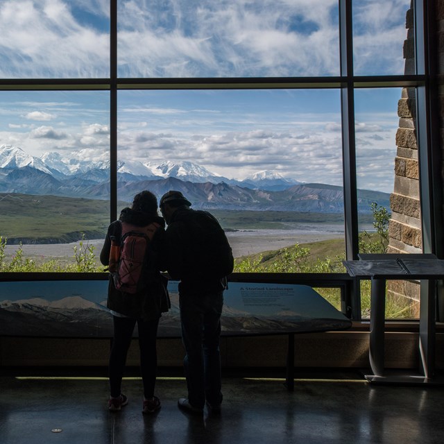 two people standing indoors in front of a window, looking out at mountains