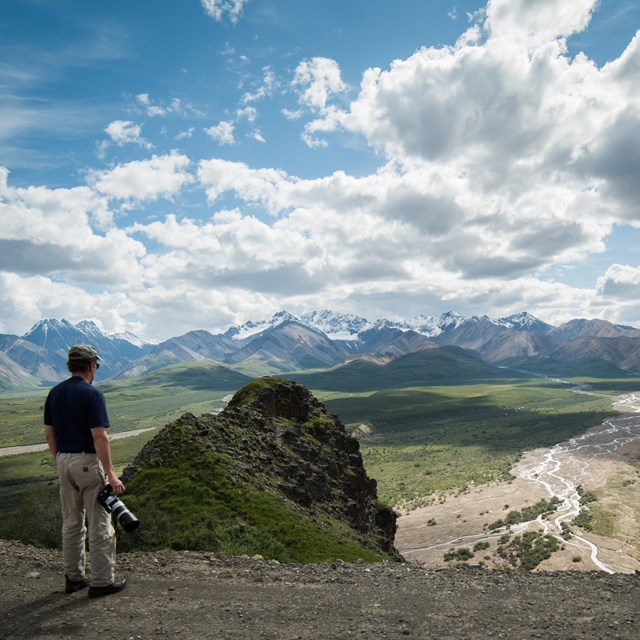 a man standing at a scenic overlook above a wide green plain leading up to distant mountains