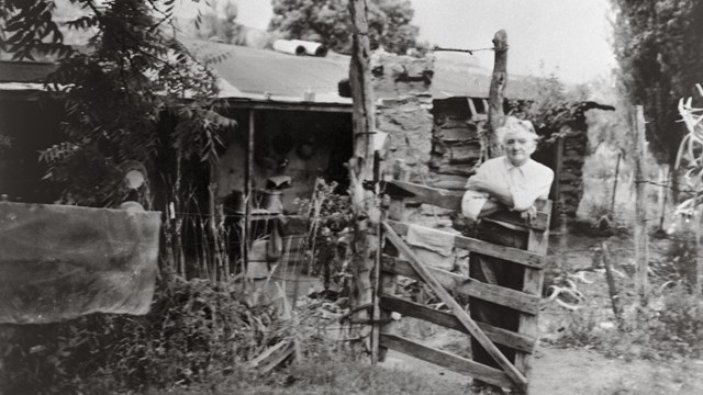 A black and white photo of a white-haired woman standing in front of a wooden gate and cabin. 