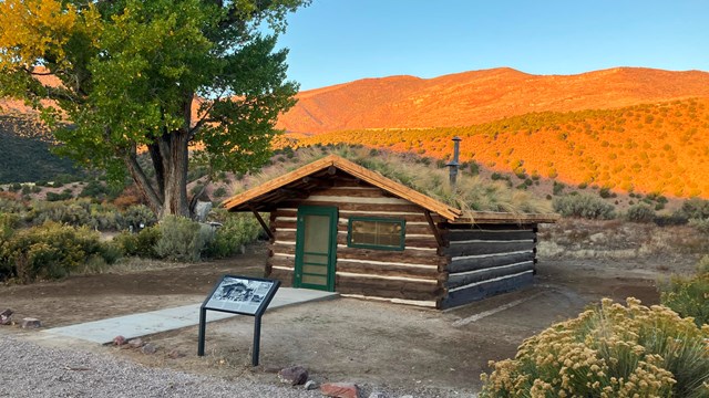 A rustic cabin with a green doorframe sitting before a large hill illuminated by the setting sun. 
