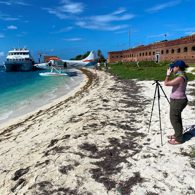 a person standing on a beach, holding a camera towards a boat, plane, and fort