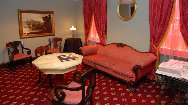 Color photo of a room furnished with red carpet, red sofa, and marble topped table.