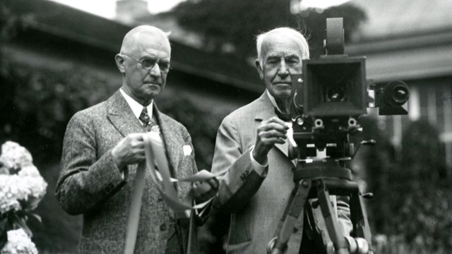 George Eastman standing next to Thomas Edison who is operating a motion picture camera.